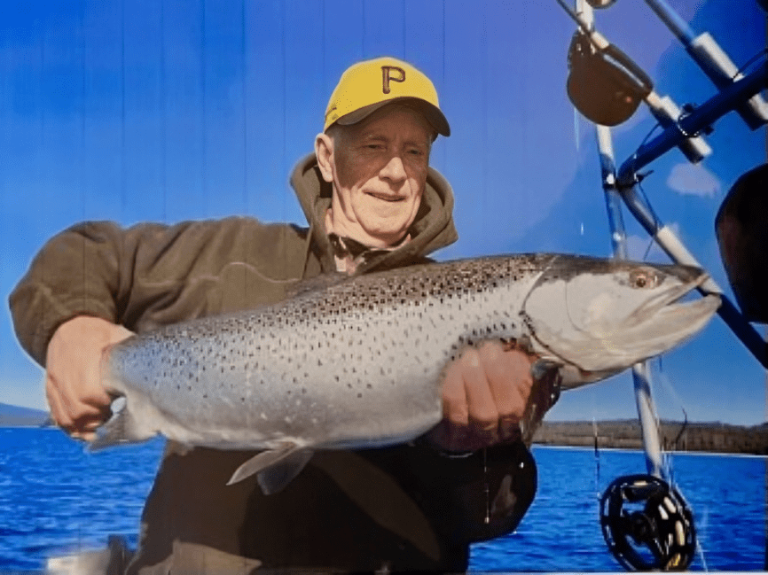 Person holding a large fish on a boat. They're wearing a yellow cap and a green jacket, with fishing equipment visible in the background.