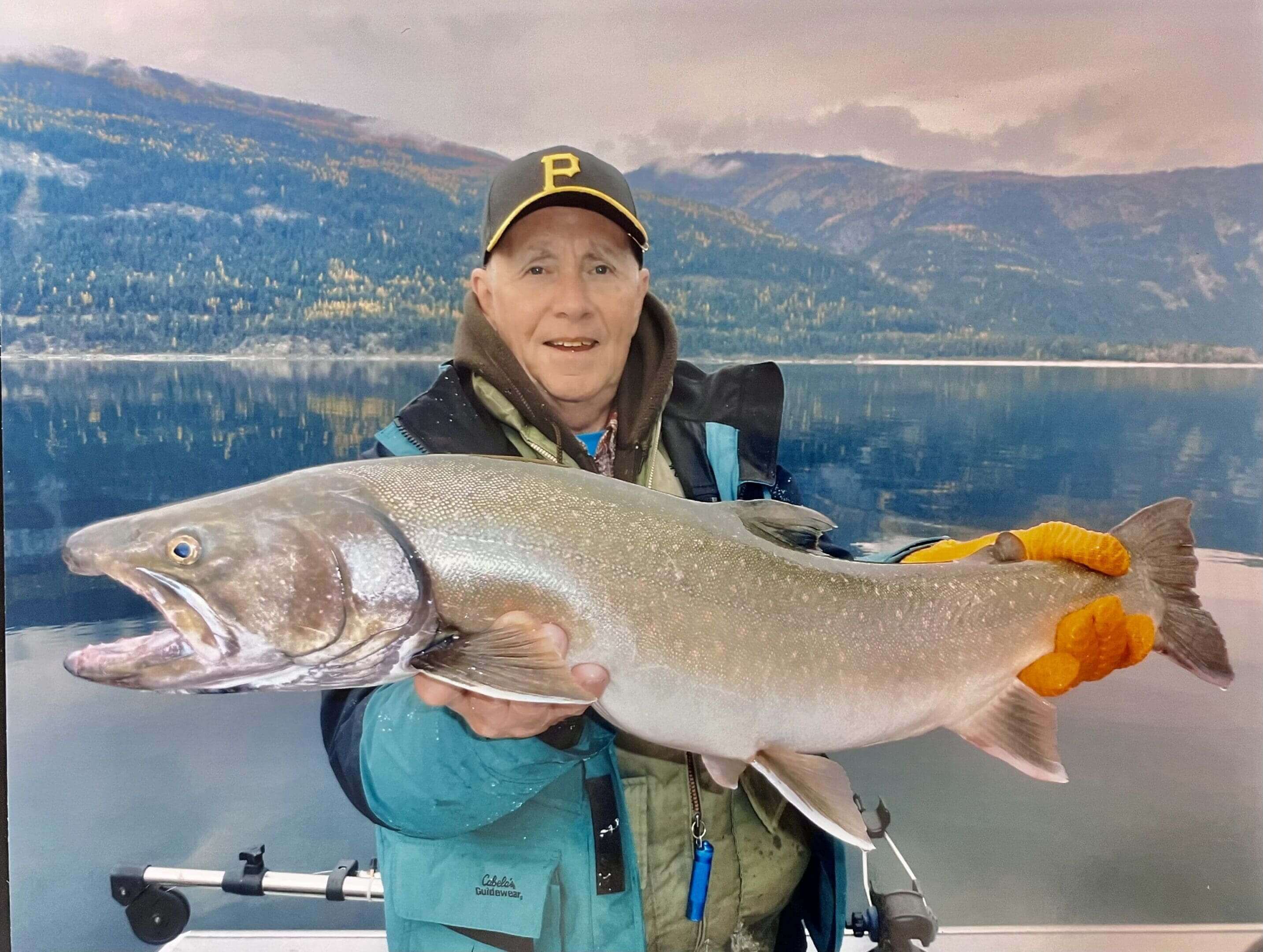 Man on a boat holding a large fish with mountains and lake in the background.