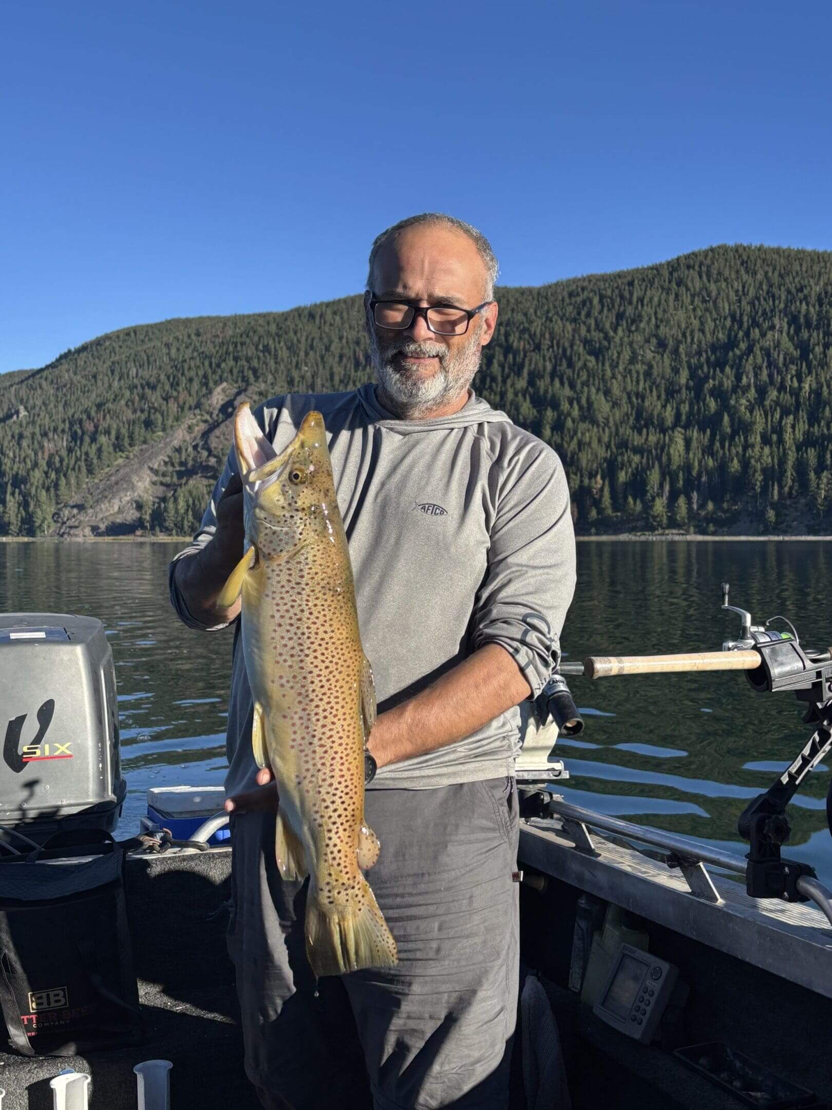 Man holding a large fish on a boat with forested hills in the background.